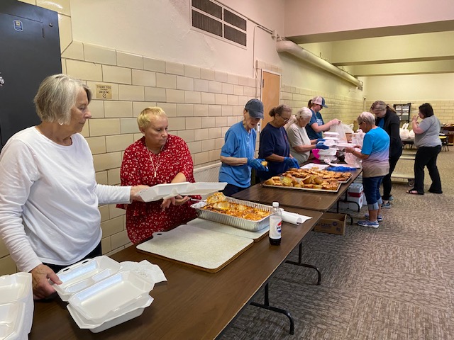 A group of people standing in front of serving tables, they are serving meals to the people in need